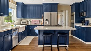 A modern kitchen with dark blue cabinetry, a central island with bar stools, stainless steel appliances, and a large window providing natural light. - Paso Wine Country Farmhouse | Pool, Spa, Game Room (Paso Robles)