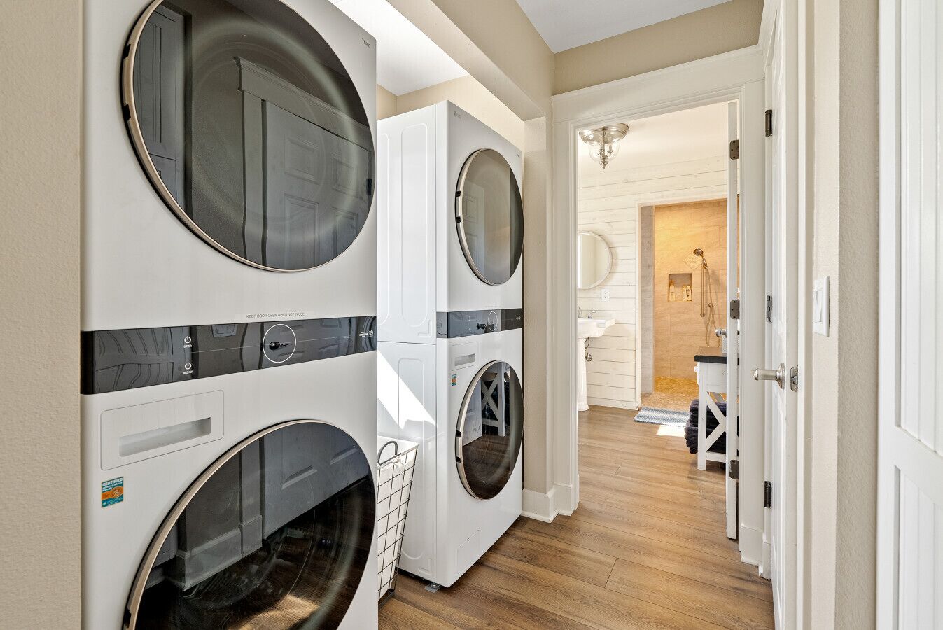 A laundry room with two front-loading washers and dryers, a wooden floor, and a hallway leading to a bathroom with a sink and mirror.