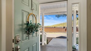 A door with a wooden wreath and greenery, leading to a covered porch with a view of a vineyard. - Paso Wine Country Farmhouse | Pool, Spa, Game Room (Paso Robles)