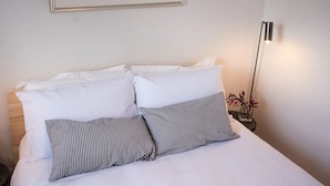 A neatly made bed with white bedding and striped pillows, a framed picture of a coastal scene on the wall, and a black pendant light. - ‘Star Cottage’ historic, cosy and walking distance to CBD/Salamanca (Sandy Bay)