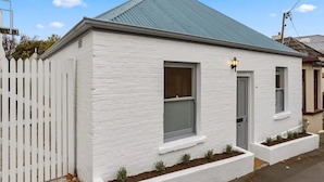 A small, single-story white building with a metal roof, a white picket fence, and a small porch with plants. - ‘Star Cottage’ historic, cosy and walking distance to CBD/Salamanca (Sandy Bay)