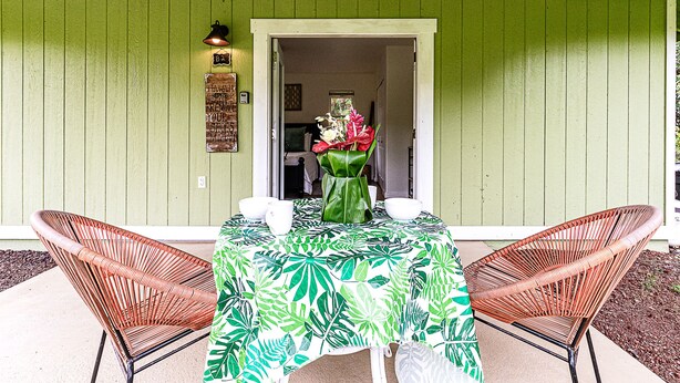 An outdoor patio with a table covered in a tropical leaf tablecloth, two wicker chairs, and a vase with flowers.