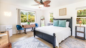 A bedroom with a bed, a desk, a chair, a ceiling fan, and large windows with a view of greenery.