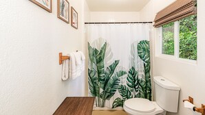 A bathroom with a toilet, a shower curtain with a tropical leaf pattern, and a window with a view of greenery.
