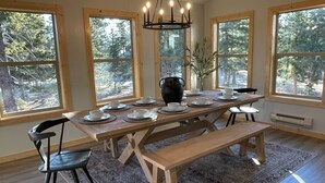 Comedor con mesa de madera dispuesta para cuatro personas, rodeado de amplias ventanas con vista a árboles.