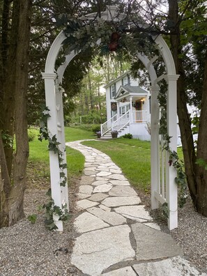 Ein Steinweg führt zu einem weißen Kletterbogen, umgeben von Bäumen und einem Haus im Hintergrund.