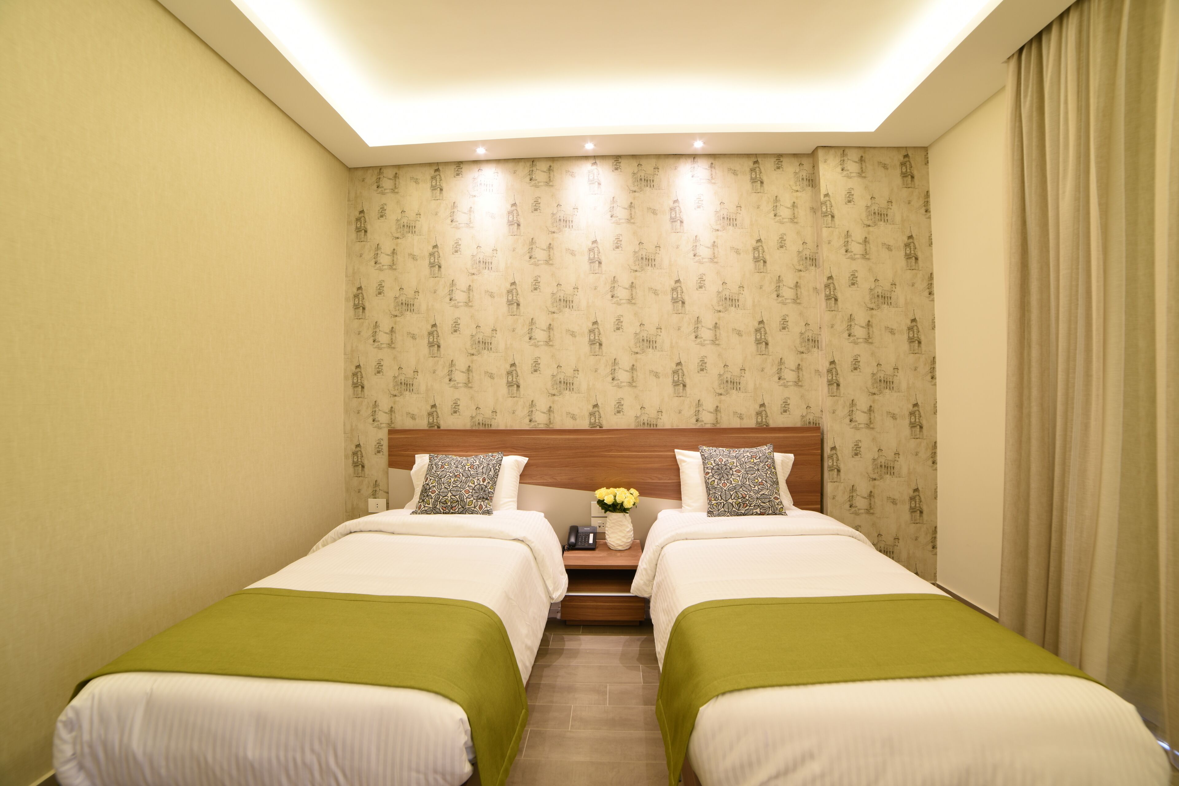 Habitación de hotel con dos camas, un cabecero de madera, una mesita de noche con un florero y un papel tapiz estampado.