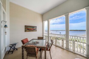 A covered balcony with a table and chairs, a painting on the wall, and a view of the water.