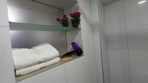 Une salle de bain aux murs recouverts de carreaux blancs, une étagère en verre sur laquelle repose une serviette blanche, un sèche-cheveux violet et deux plantes en pot ornées de fleurs roses.