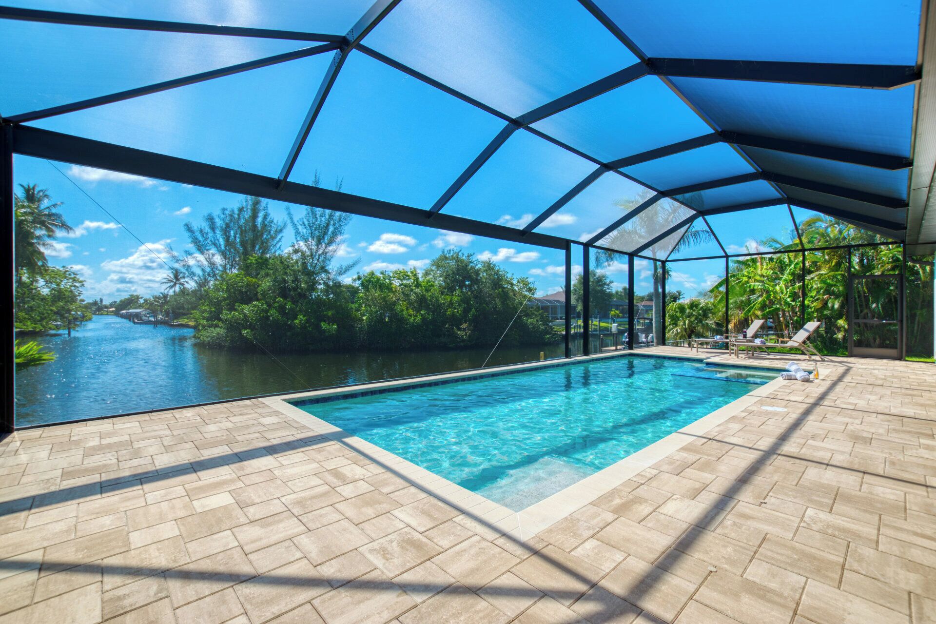 A swimming pool with a clear view of a river and lush greenery, surrounded by a covered area with tiled flooring and lounge chairs.