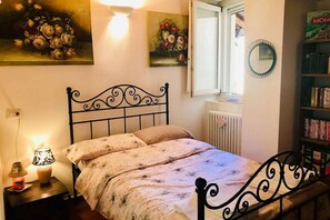 Un dormitorio con una cama, una lámpara de noche, un radiador, una ventana y una estantería.