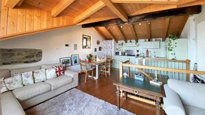 A cozy living room with a wooden ceiling, a stone wall, a dining area with a table and chairs, and a kitchen with light-colored cabinets. - Cà Milla Argegno apartment (Argegno)