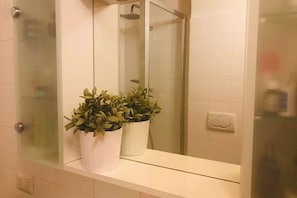 Un baño con un lavamanos blanco, dos plantas en macetas, un box de ducha de cristal y una pared con azulejos blancos.