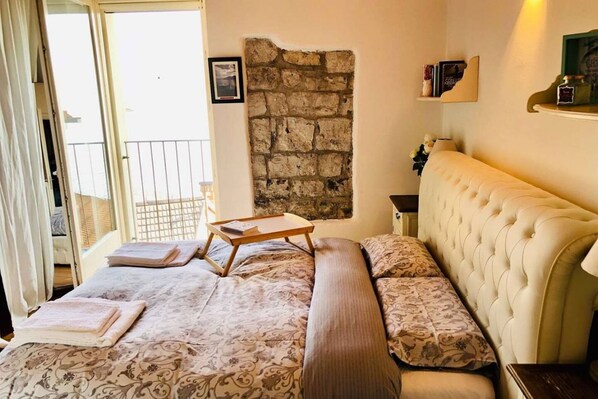 Un dormitorio con cama, mesita de noche, pared de piedra, balcón y ventana.