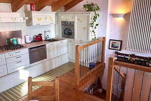 Una cocina con armarios blancos, una estufa y un lavamanos. Se observa una escalera de madera con barandilla.