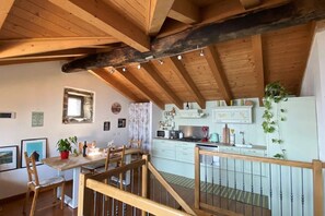 A rustic kitchen with wooden beams, a dining area with chairs, and a staircase leading to an upper level.
