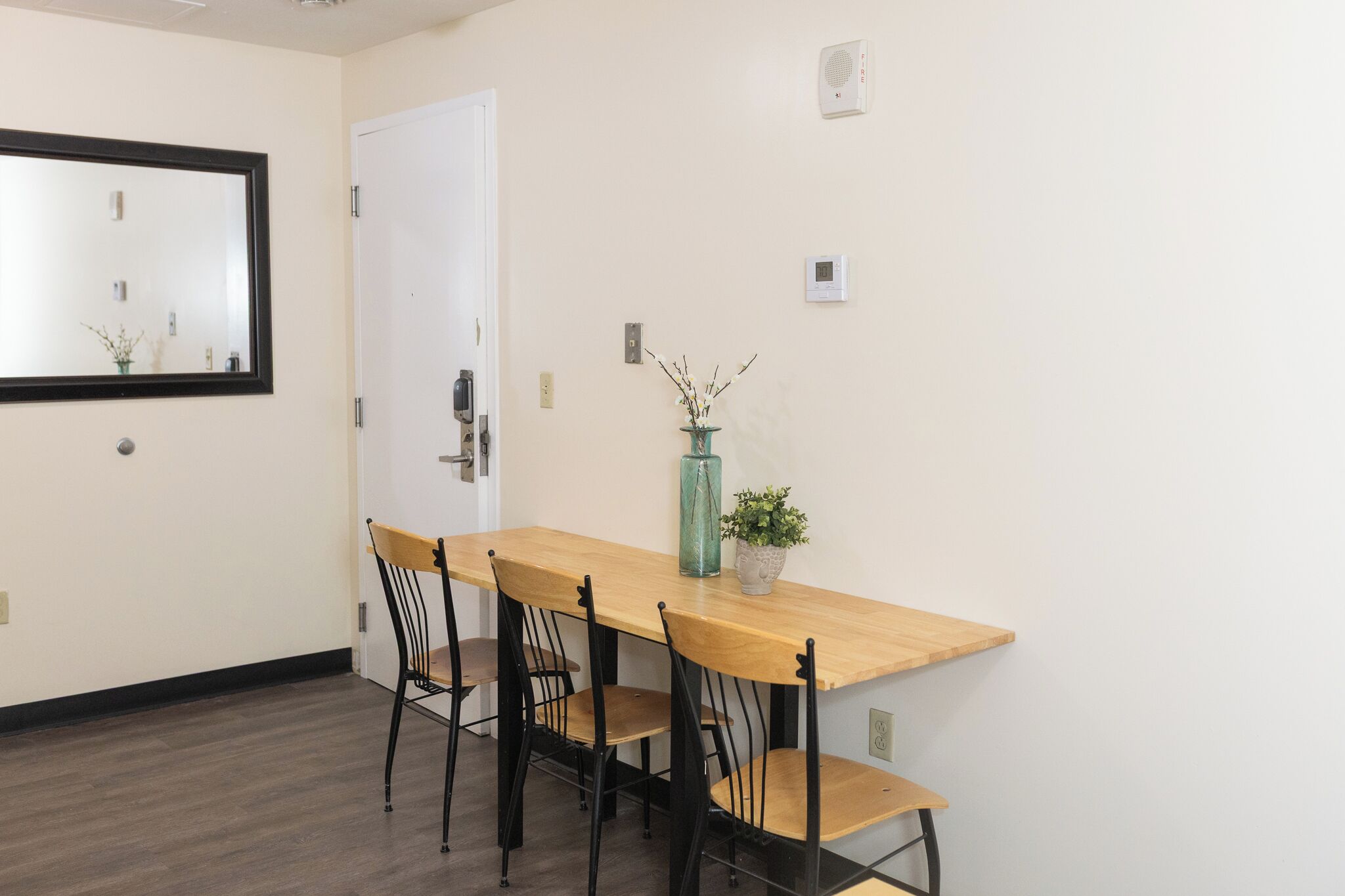 A dining area with a wooden table, four chairs, a vase with branches, and a door.