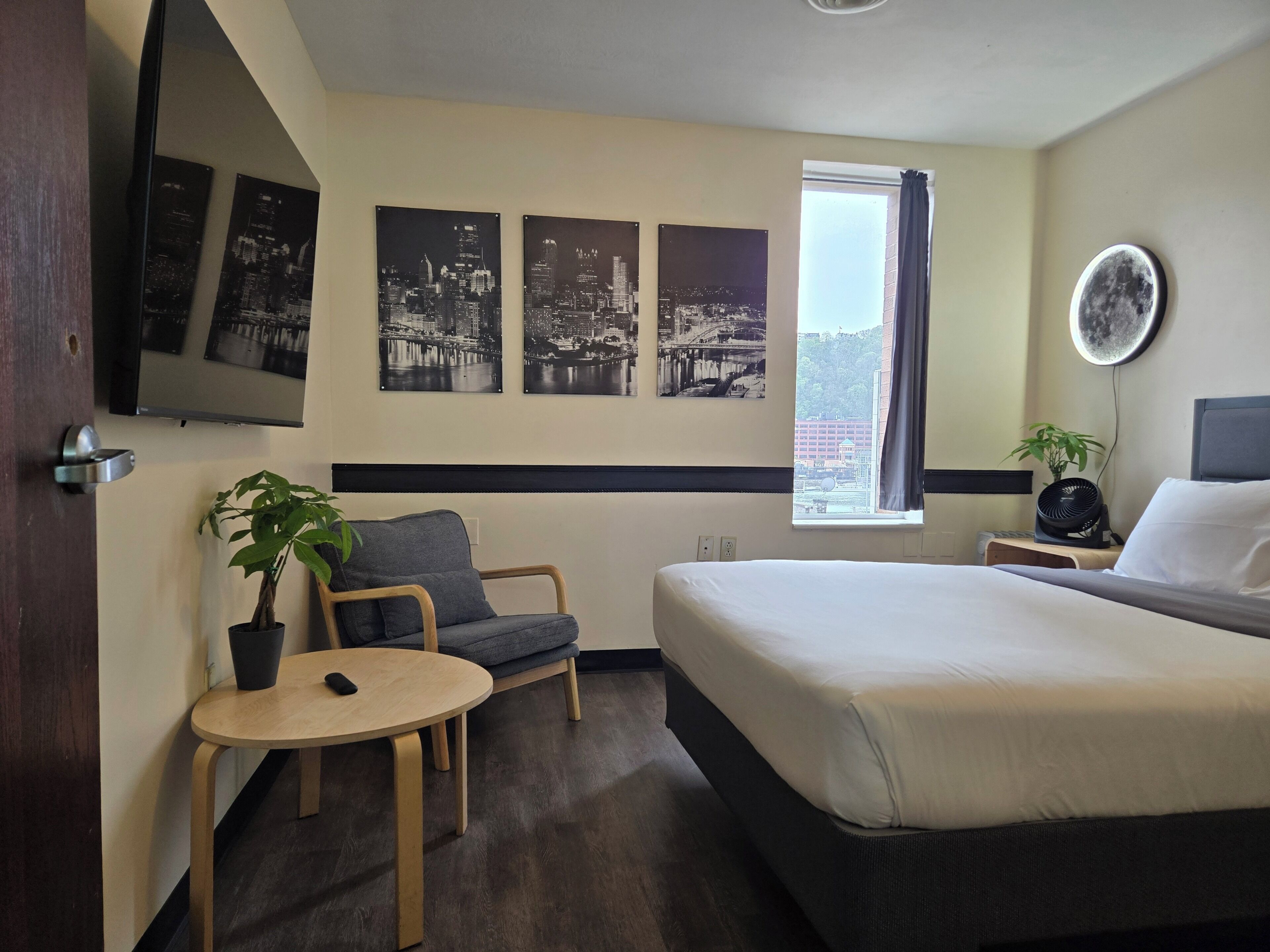 Habitación de hotel con cama, una silla, una mesita con una planta, televisión de pared y fotografías del paisaje urbano.