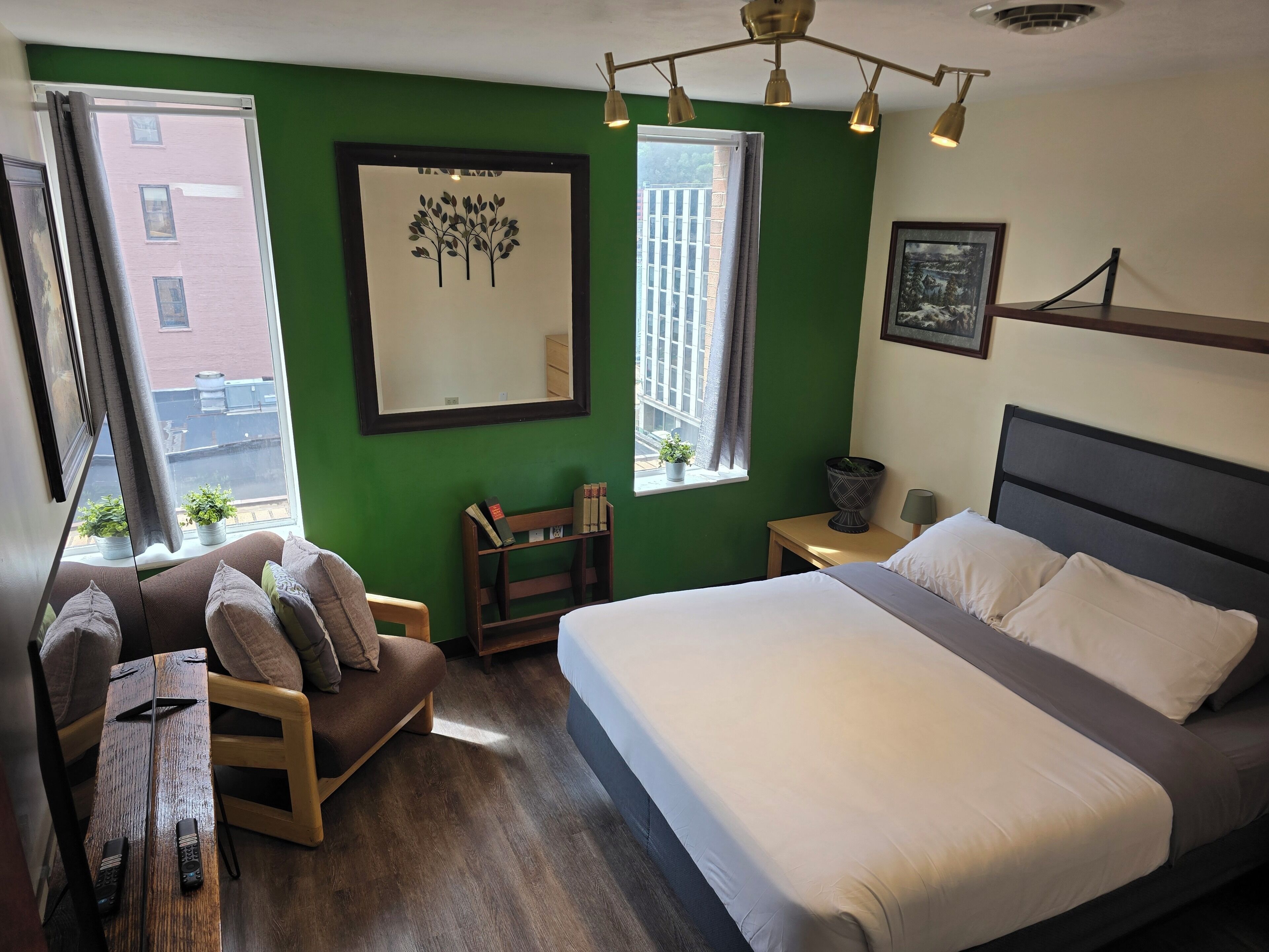 Ein Schlafzimmer mit Bett, Sessel, kleinem Tisch, Nachttisch, Spiegel und zwei Fenstern.