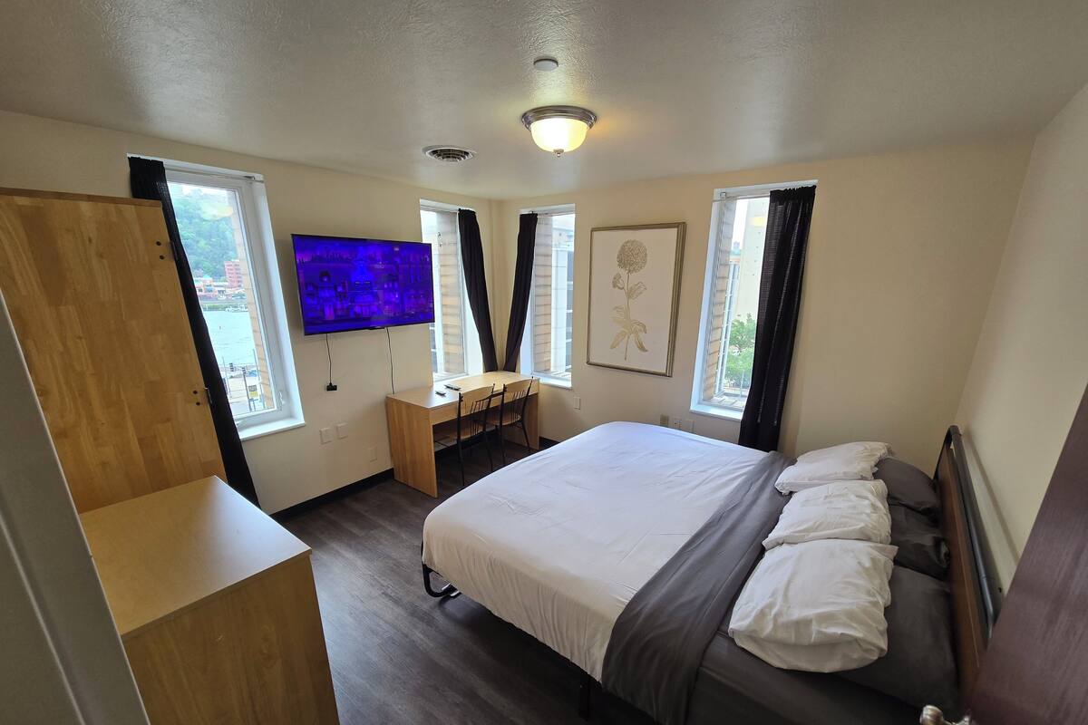 A bedroom with a bed, desk, chair, TV, windows, and a door.