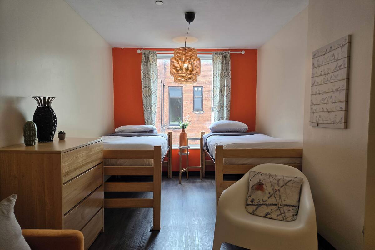 A room with two beds, a desk, a chair, and a hanging light fixture.
