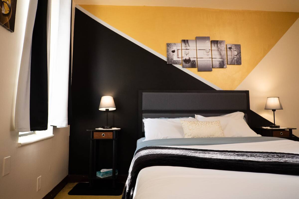 A hotel room with a bed, two bedside tables with lamps, a wall with a geometric artwork, and a window with curtains.