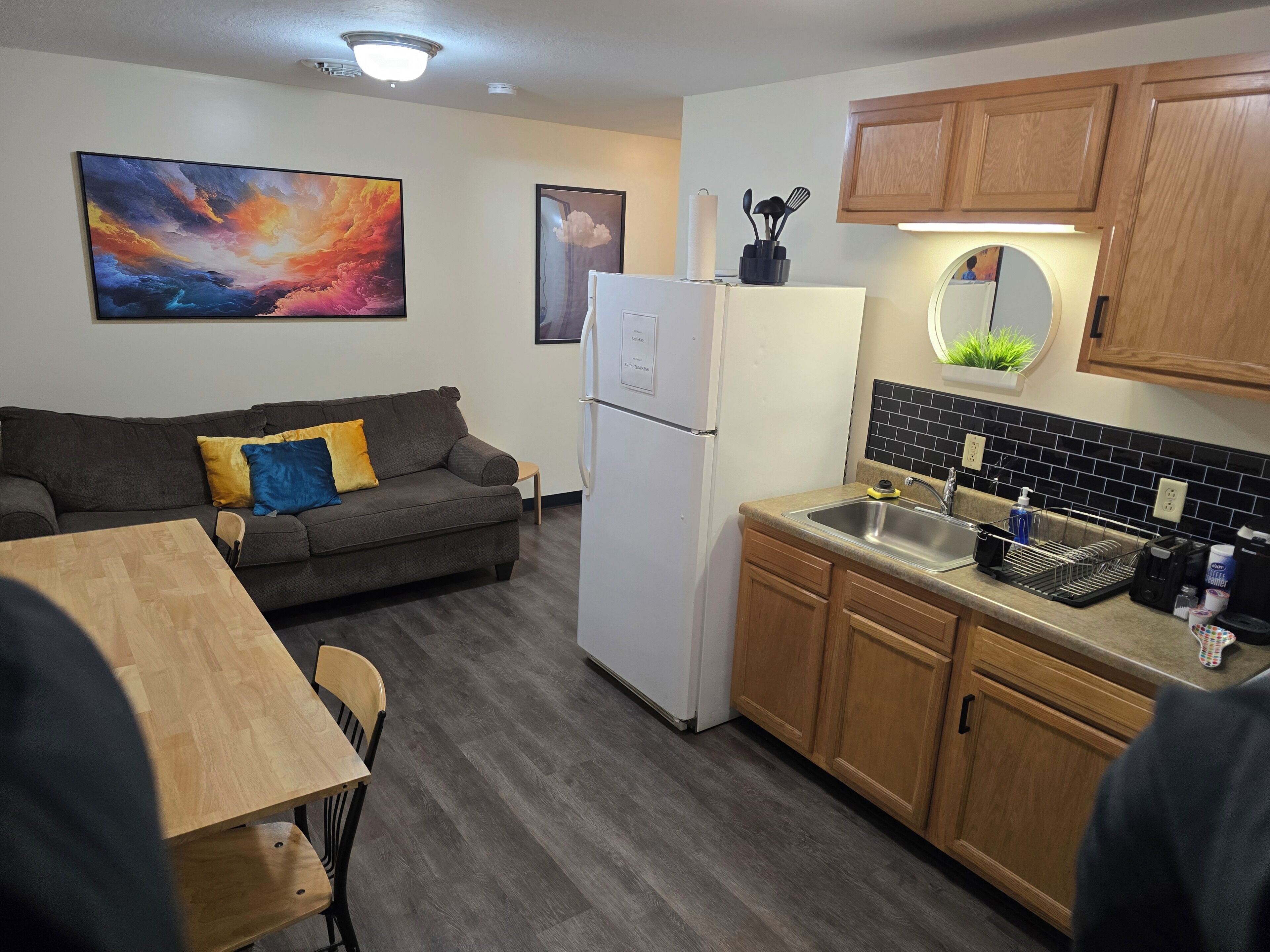 Cocina compacta con refrigerador, armarios de madera, un lavamanos y una zona de comedor con mesa y sillas.