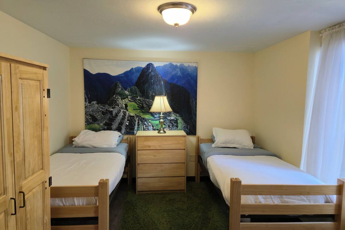 A room with two single beds, a wooden dresser, a lamp, and a large painting of Machu Picchu on the wall.