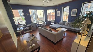 A modern living room with a sofa, a coffee table, a dining area with a small table and chairs, and large windows offering city views. - Stay Pittsburgh (Pittsburgh)