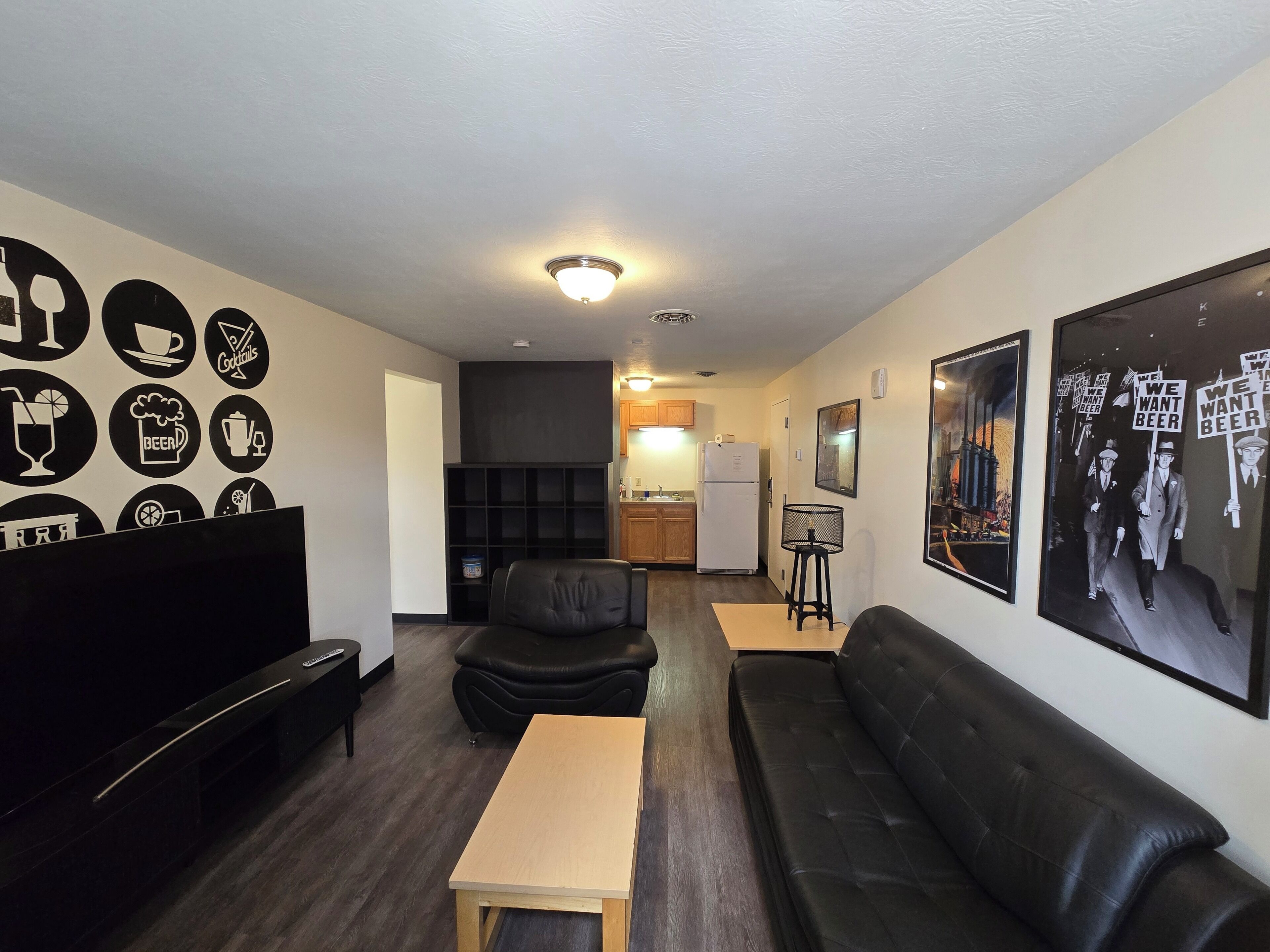 A modern living room with a black leather sofa, a wooden coffee table, and a black armchair. There is a kitchen area in the background with wooden cabinets and a refrigerator.