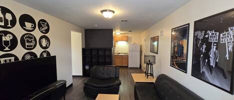 A modern living room with a black leather sofa, a wooden coffee table, and a black armchair. There is a kitchen area in the background with wooden cabinets and a refrigerator.