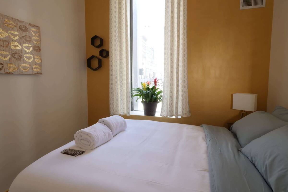 A hotel room with a neatly made bed, a window with curtains, a potted plant, and a wall-mounted artwork.