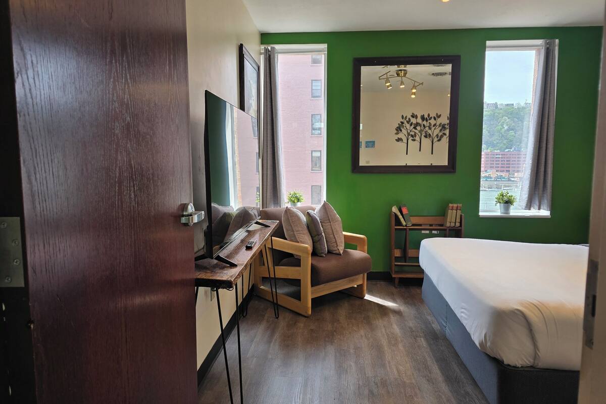 A hotel room with a bed, a desk, a chair, a TV, and a window with a view of buildings.