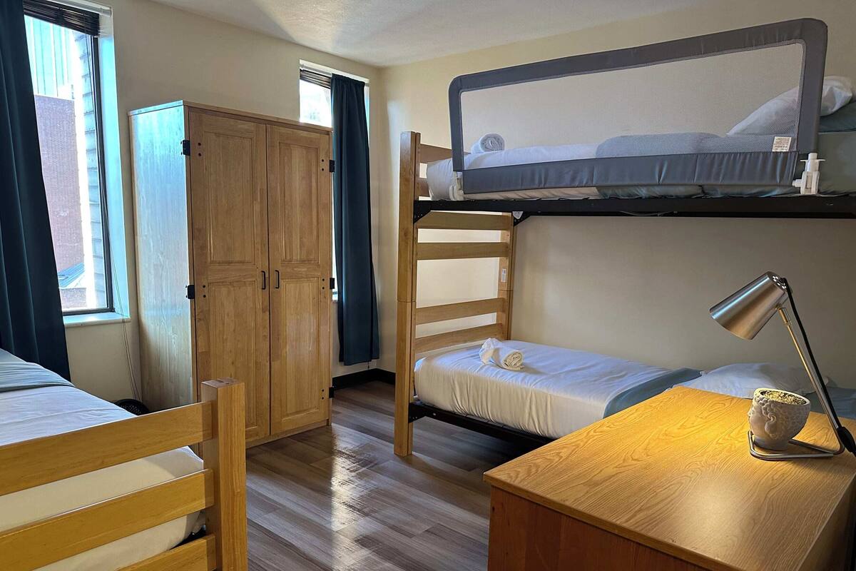 A compact room with a bunk bed, a single bed, a wooden wardrobe, and a desk with a lamp.