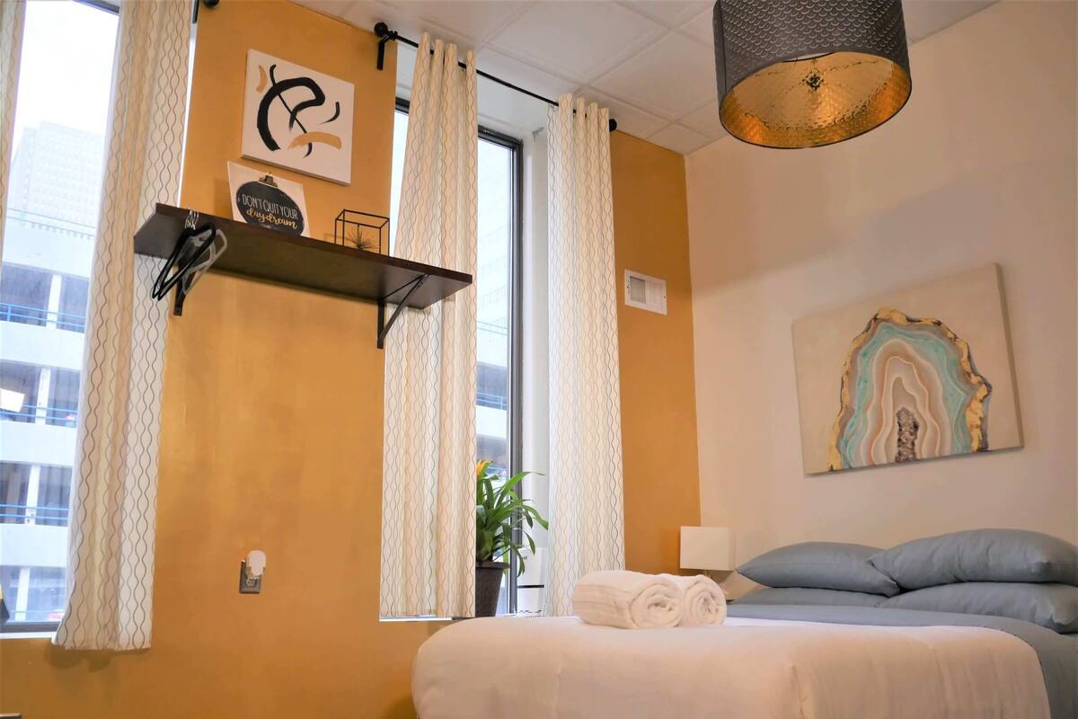 A hotel room with a bed, a shelf with toiletries, a painting, and a window with curtains.