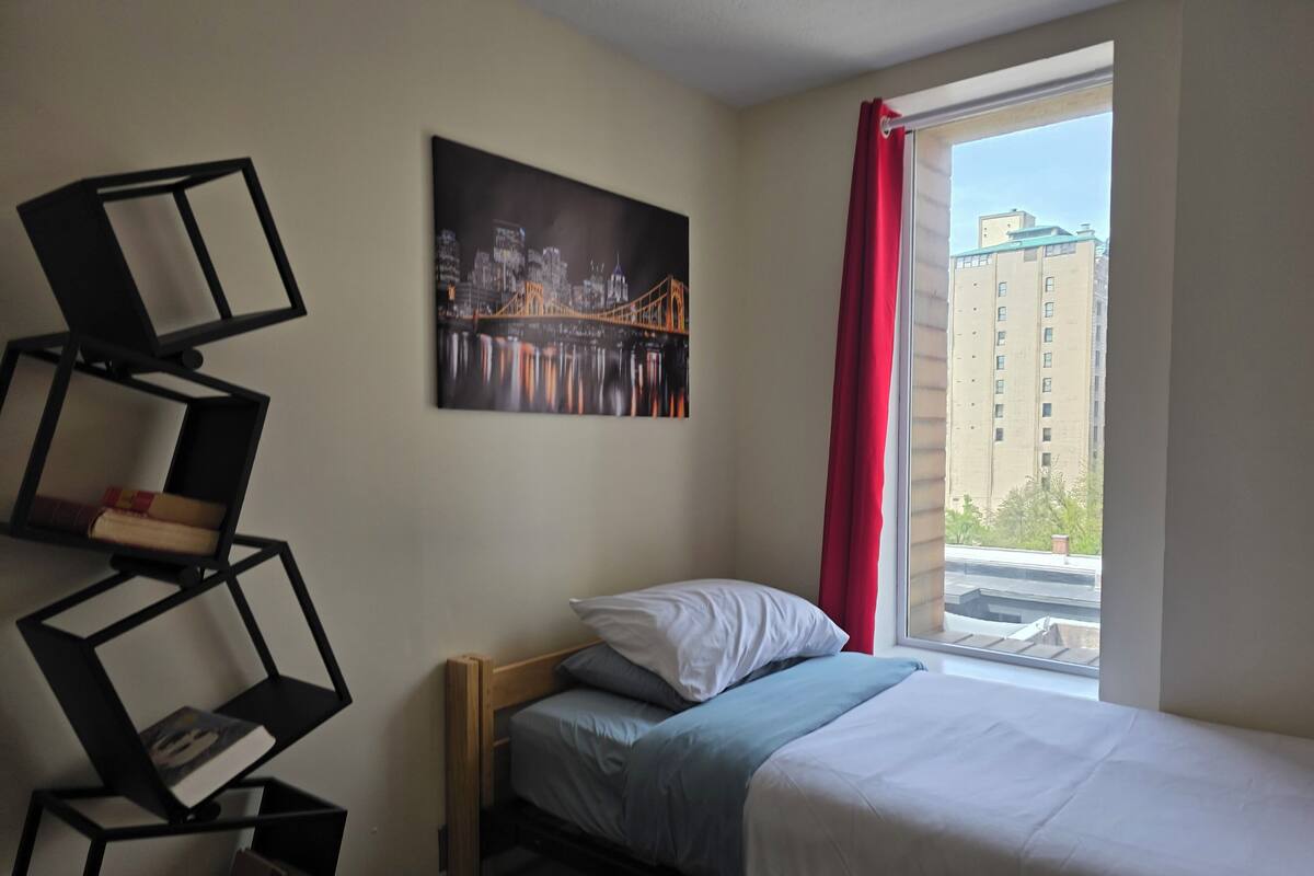 A bedroom with a bed, a window with a red curtain, a cityscape painting, and a geometric bookshelf.