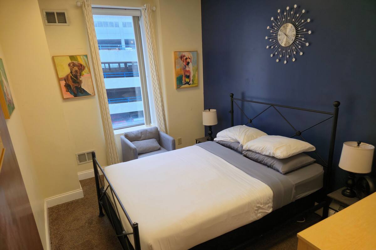 A bedroom with a bed, two bedside lamps, a wall clock, and a window with curtains.