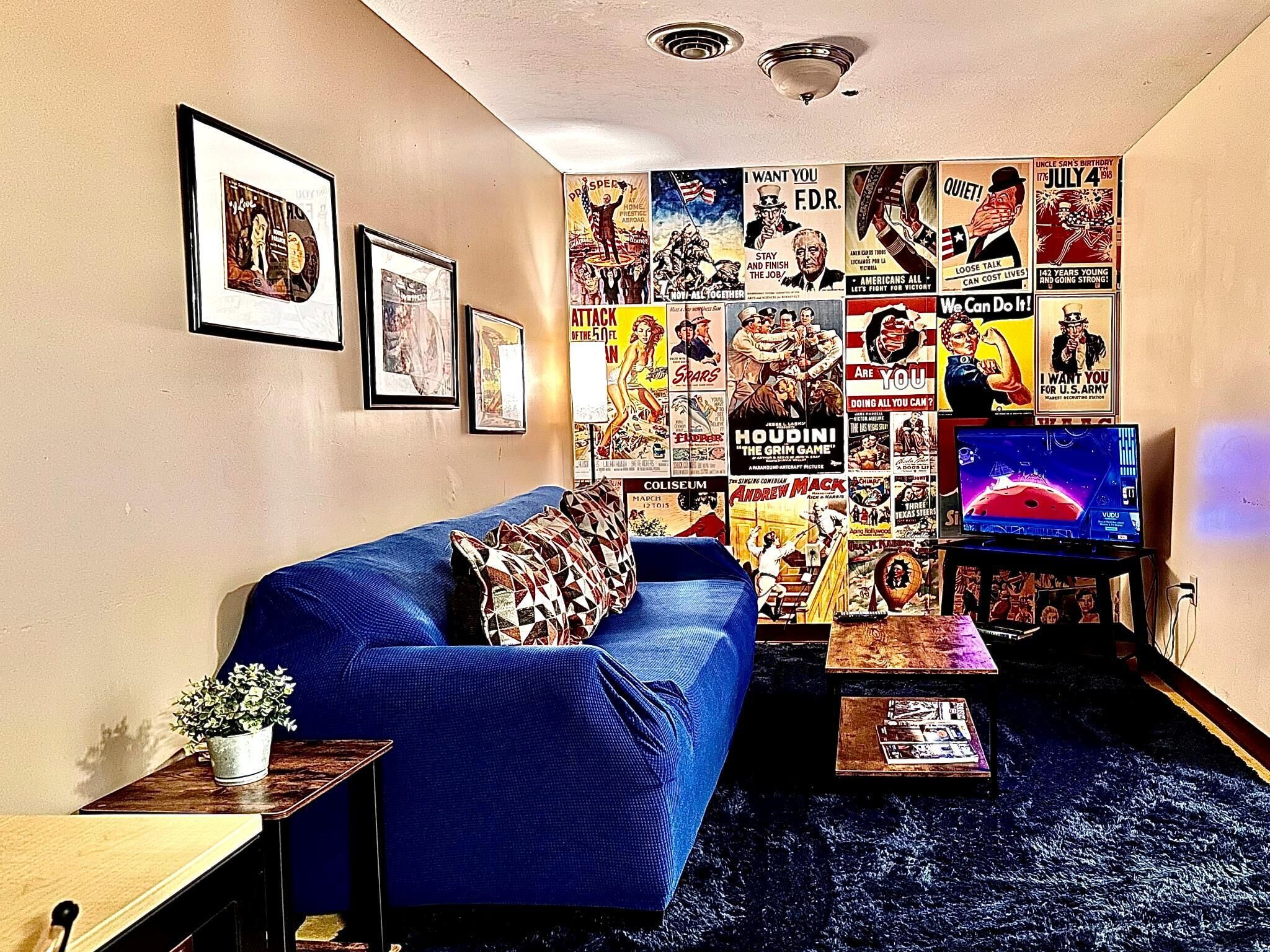 A living room with a blue sofa, a coffee table, and a television. The walls are decorated with various posters and pictures.