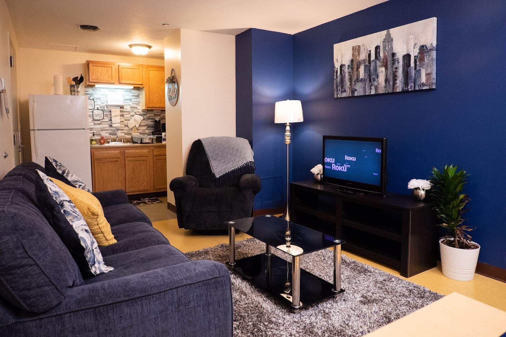 A living room with a dark blue wall, a flat-screen TV, a sofa, a chair, a lamp, and a small potted plant.