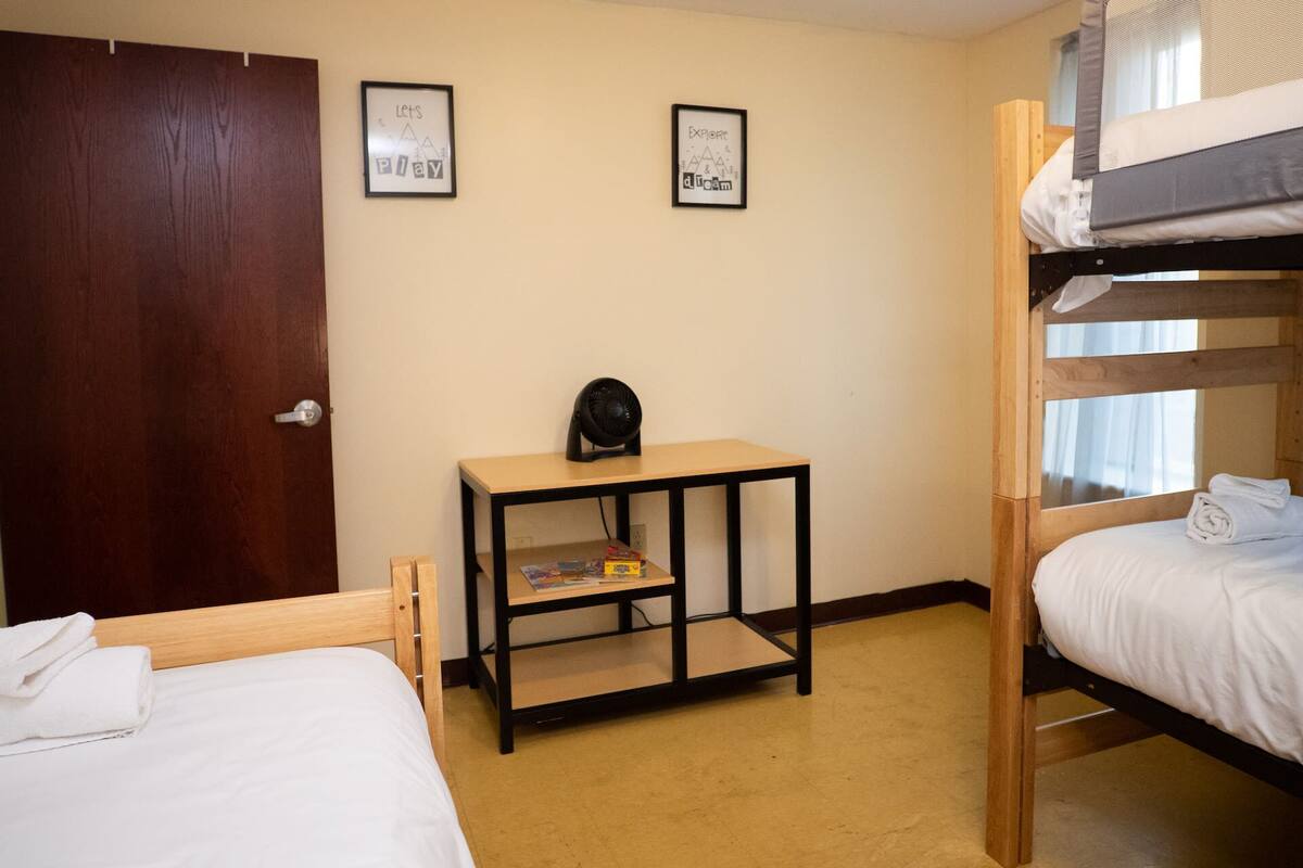 A bunk bed room with a wooden door, two framed pictures on the wall, a small table with a fan, and a window with curtains.