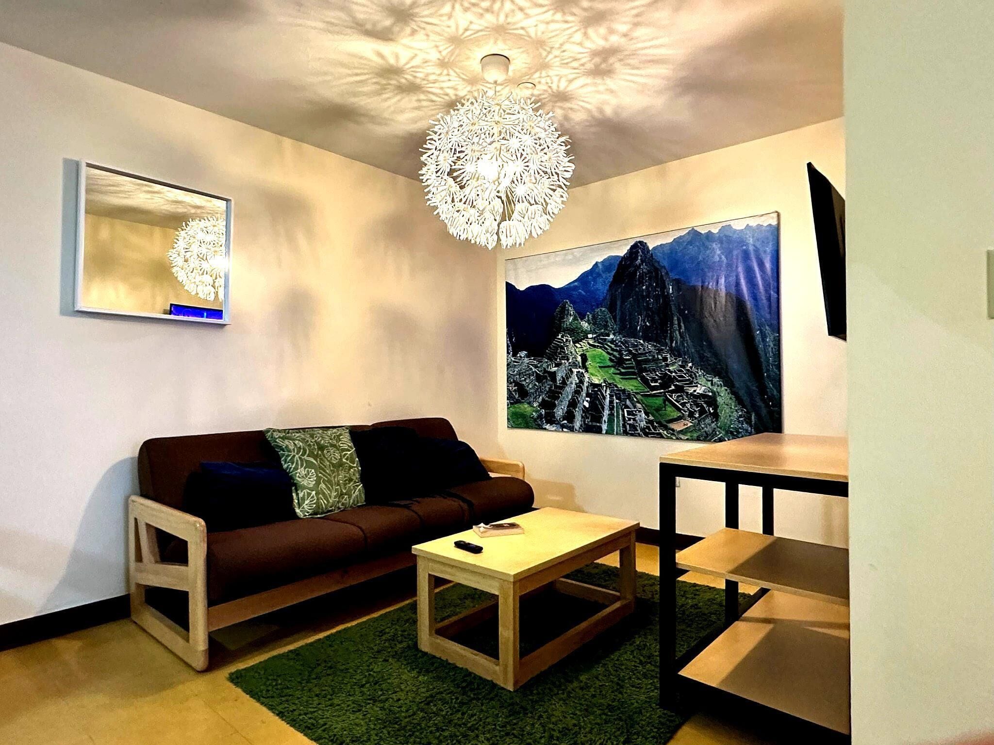 A modern living room with a brown sofa, a small wooden coffee table, a green rug, a chandelier, and a large painting of Machu Picchu.
