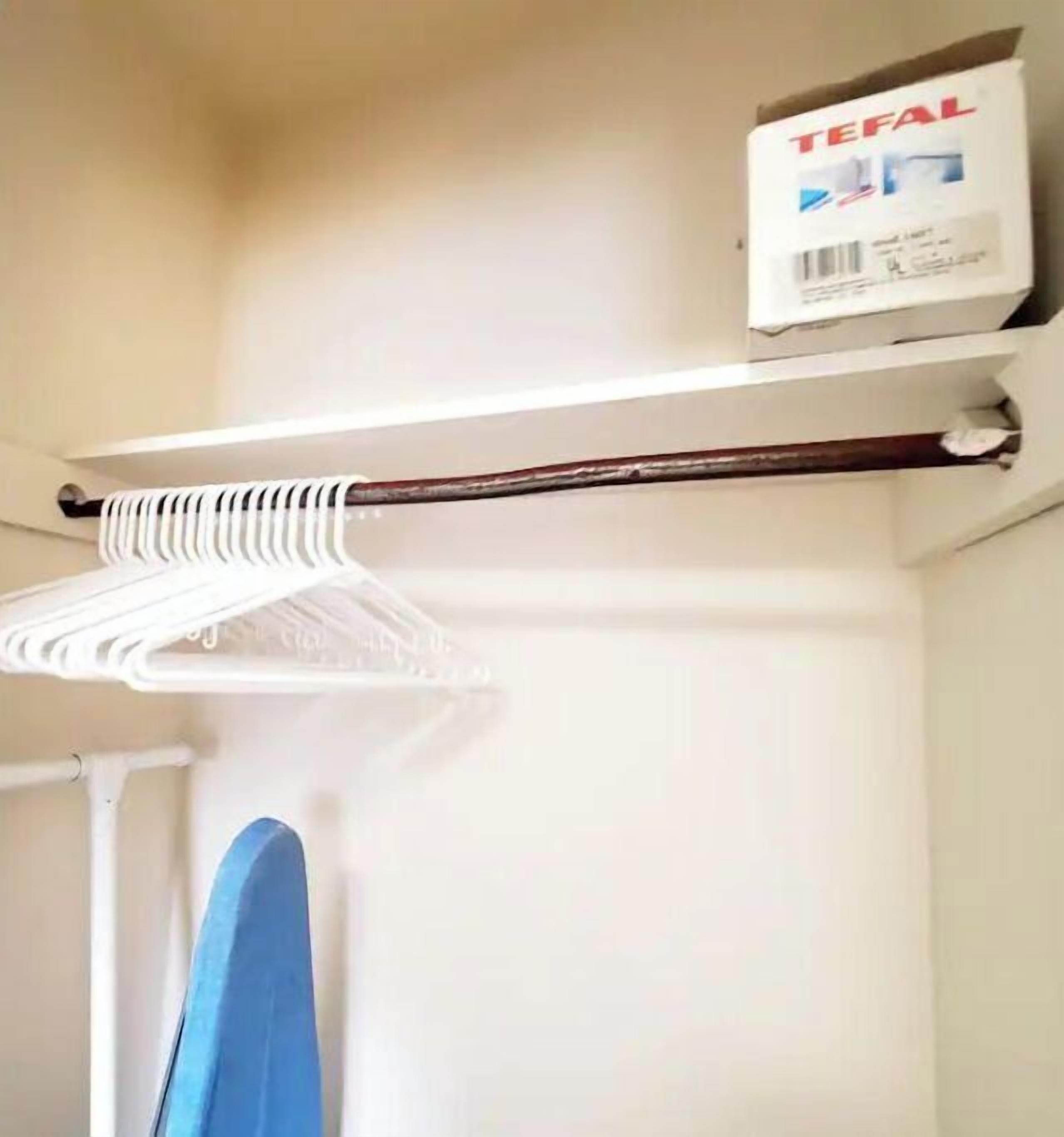 A white wall with a white shelf holding a box labeled "TEFAL" and a wooden rod with a white cloth hanging from it.