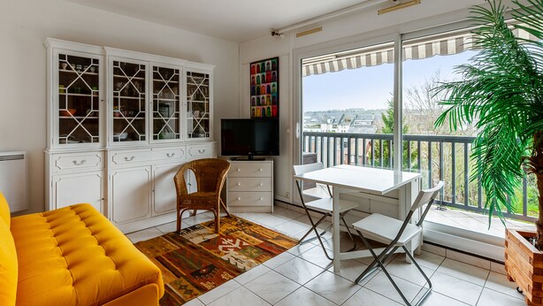 A bright living room with a TV, white cabinets, a wicker chair, a yellow sofa, and a view of the city through large windows. - Studio Conac - Welkeys (Trouville-sur-Mer)