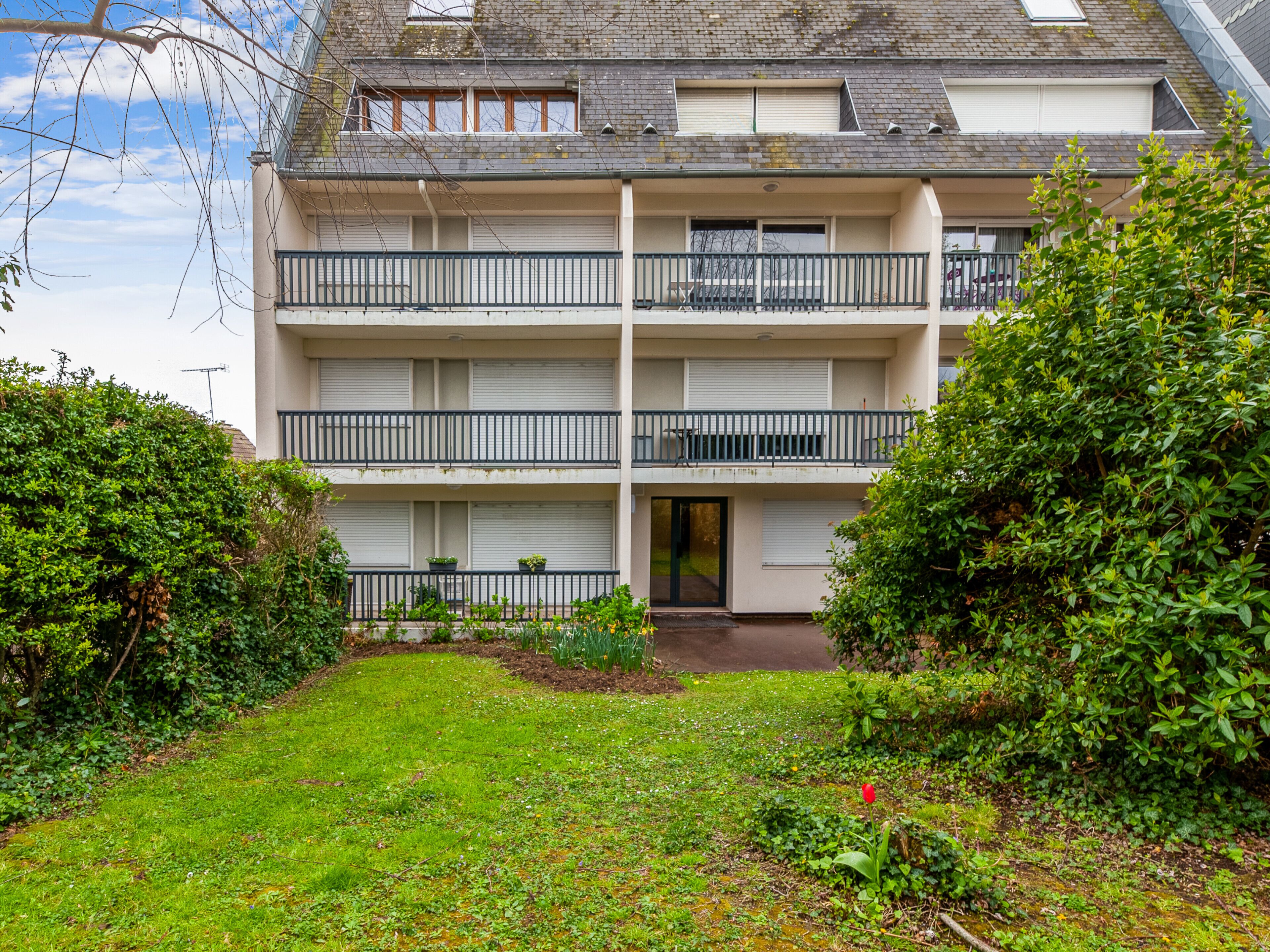 A multi-story residential building with balconies, a front door, and a small garden area with grass and plants.