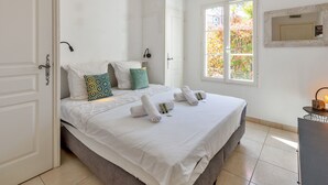 A hotel room with a large bed, white bedding, a window with a view of trees, a bedside table with a lamp, and a door with a keyhole. - Appartement du Brost - Résidence le clôt des Sarments - Welkeys (La Croix-Valmer)