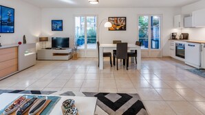 A modern living room with a TV, seating, and a dining area. There are windows and a kitchen visible. - Appartement du Brost - Résidence le clôt des Sarments - Welkeys (La Croix-Valmer)