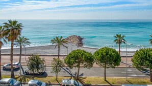 A coastal road with parked cars, palm trees, and a rocky outcrop by the sea. - Appartement Rivage - Welkeys (Cagnes-sur-Mer)