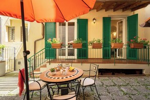 Un patio al aire libre con una mesa redonda, sillas y un gran paraguas naranja. Hay plantas en macetas en el alféizar de la ventana y una escalera que lleva a un nivel superior.