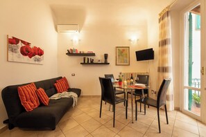 Una sala de estar con un sofá negro, cojines rojos, una mesa de comedor con sillas, un televisor montado en la pared y una ventana con cortinas.