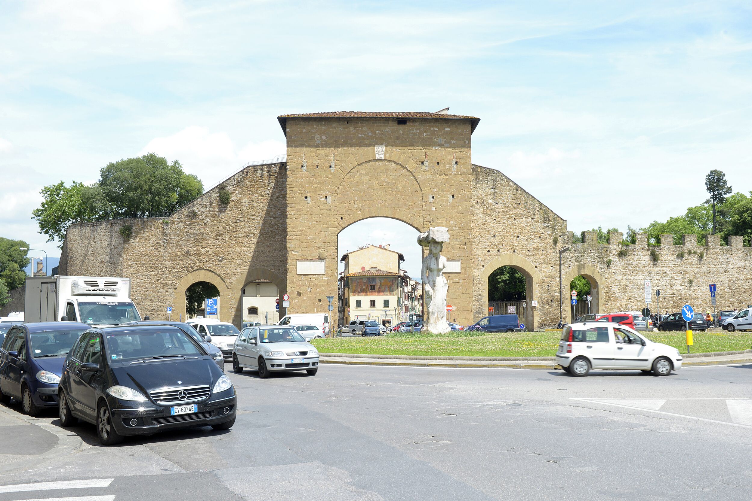 Un arco de piedra histórica con una estatua en primer plano, rodeado de coches aparcados y un cielo despejado.